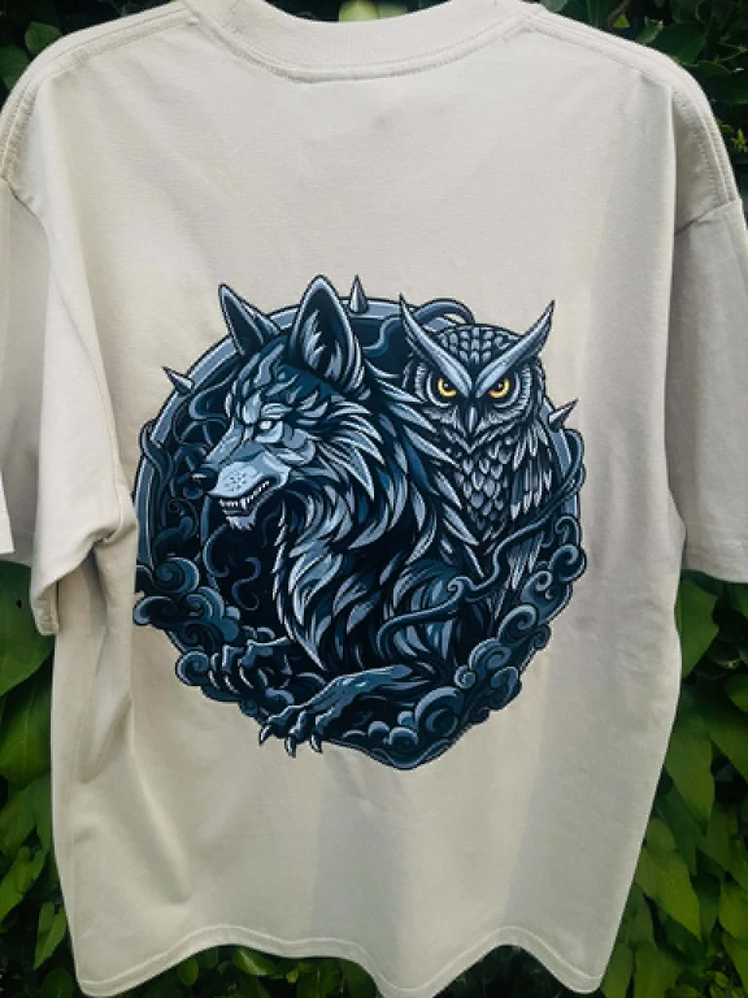 An light green short-sleeve t-shirt featuring a large graphic print of a blue wolf and owl in a circular design, set against a background of green foliage.